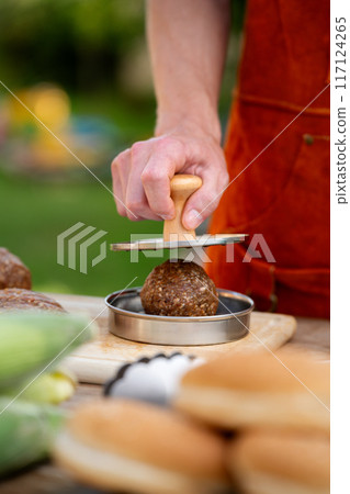 Man in apron preparing meat for hamburgers, standing outdoors. Using burger patty press to shape groud meat. 117124265