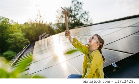 Cute girl on roof with solar panels, holding model of wind turbine. Rooftop solar or photovoltaic system. Sustainable future for next generation. 117124283