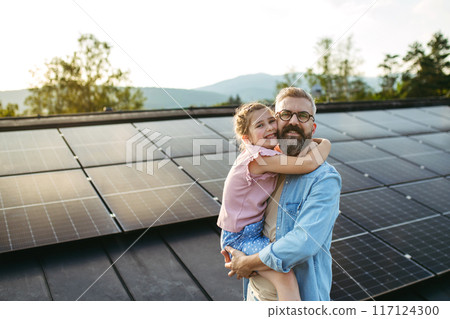 Father with girl on roof with solar panels, hugging, looking at camera. Rooftop solar or photovoltaic system. Sustainable future for next generation. 117124300