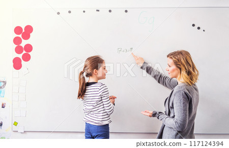 Hardworking teacher looking at young schoolgirl in classroom. Girl student writing on whiteboard. Hardworking teacher looking at young schoolgirl in classroom. Girl student writing on whiteboard. 117124394