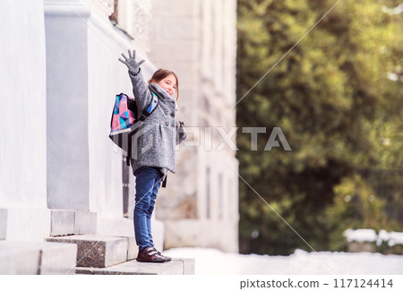 Portrait of schoolgirl in coat in front of school building, waiting for parents. Student girl with bag after day in elementary school. Portrait of schoolgirl in coat in front of school building, waiting for parents. Student girl with bag after day in elementary school. 117124414