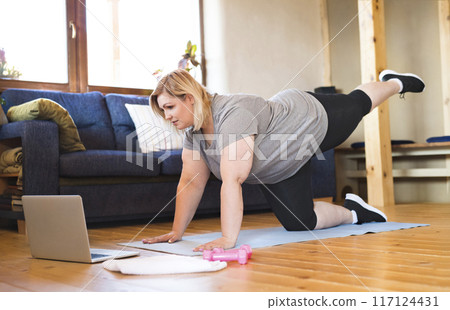 Online fitness training for overweight woman exercising at home, looking at laptop. Workout in living room. 117124431