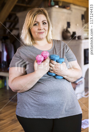 Overweight woman exercising at home, holding blue and pink dumbbells. Workout in living room. Overweight woman exercising at home, holding blue and pink dumbbells. Workout in living room. 117124433