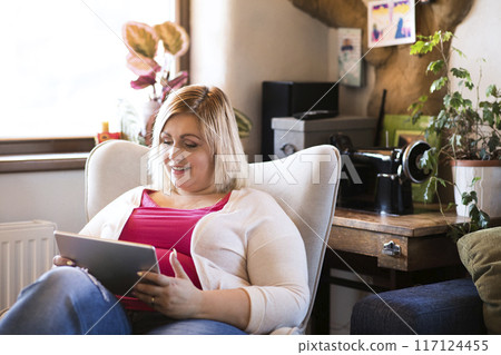 Portrait of beautiful young overweight woman at home, watching tv show on tablet, sitting in armchair. 117124455