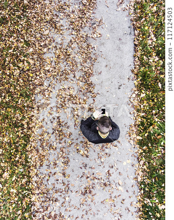 Aerial view of a runner standing autumn park on path with fallen leaves, holding smartphone, checking training performance on smartwatch. Morning running training. Aerial view of a runner standing autumn park on path with fallen leaves, holding smartphone, checking training performance on smartwatch. Morning running training. 117124503