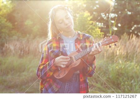 Girl playing ukulele, sunset music session, summer harmony. girl strumming guitar, summer vibes, outdoor music therapy. Blurred 117125267