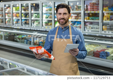 Supermarket worker holding frozen food package and using digital tablet in front of refrigerated section. Smiling male employee in apron managing inventory. Modern grocery store environment. Supermarket worker holding frozen food package and using digital tablet in front of refrigerated section. Smiling male employee in apron managing inventory. Modern grocery store environment. 117129753