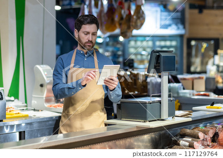 Male grocery store worker using tablet at checkout counter. Worker wearing apron, standing in modern supermarket, checking inventory and prices on device. Male grocery store worker using tablet at checkout counter. Worker wearing apron, standing in modern supermarket, checking inventory and prices on device. 117129764