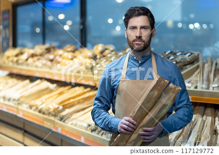 Baker holding fresh baguettes in front of supermarket bread section. Wearing apron, standing in bakery aisle with baked goods in background. Profession, bread, bakery, grocery store concept. Baker holding fresh baguettes in front of supermarket bread section. Wearing apron, standing in bakery aisle with baked goods in background. Profession, bread, bakery, grocery store concept. 117129772