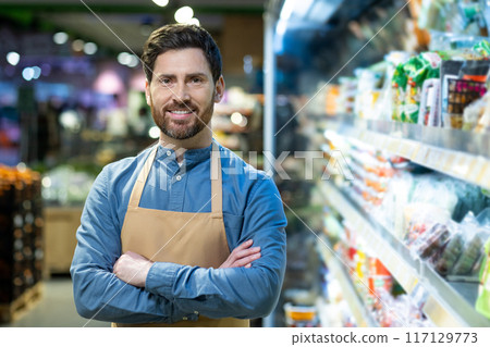 Confident male grocery store employee standing with arms crossed in supermarket aisle. Worker wears apron and smiles at camera surrounded by various goods. Professionalism and retail work concept. 117129773