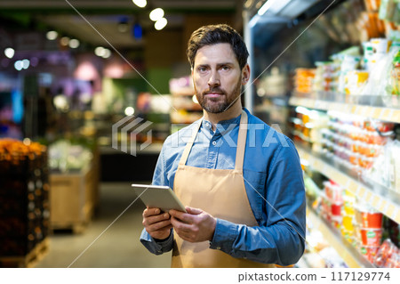 Supermarket worker using tablet in grocery store. Person wearing apron and holding tablet, performing work duties. Shelves stocked with products visible in background. Supermarket worker using tablet in grocery store. Person wearing apron and holding tablet, performing work duties. Shelves stocked with products visible in background. 117129774