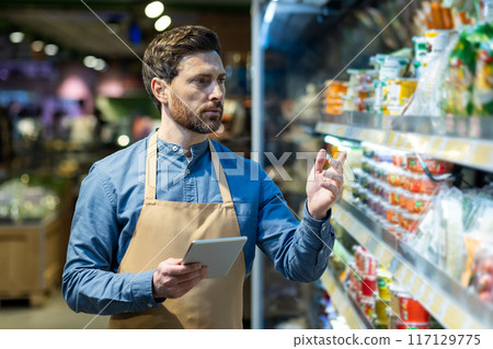Supermarket employee checking products on shelves while holding tablet. Worker managing inventory and ensuring product availability. Focused on quality control and organization in grocery store. Supermarket employee checking products on shelves while holding tablet. Worker managing inventory and ensuring product availability. Focused on quality control and organization in grocery store. 117129775
