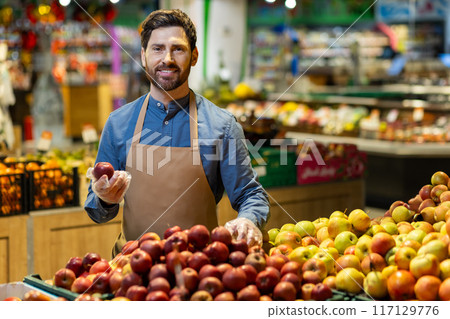 Smiling shopkeeper holding apple at fruit stand in grocery store. Man in apron displaying fresh produce, engaging with customers, promoting healthy eating and organic food Smiling shopkeeper holding apple at fruit stand in grocery store. Man in apron displaying fresh produce, engaging with customers, promoting healthy eating and organic food 117129776