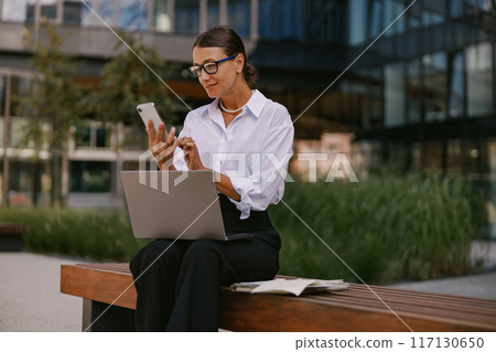 A Professional Woman Engaged in Her Work Outdoors Using a Laptop and a Mobile Phone A Professional Woman Engaged in Her Work Outdoors Using a Laptop and a Mobile Phone 117130650