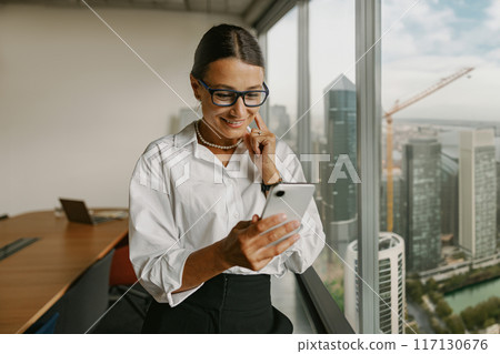 A Professional Woman Engaged with Her Smartphone While Present in a Modern Office Environment A Professional Woman Engaged with Her Smartphone While Present in a Modern Office Environment 117130676