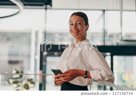 Smiling and Confident Professional Woman Positioned in a Modern Office Setting, Holding a Smartphone Smiling and Confident Professional Woman Positioned in a Modern Office Setting, Holding a Smartphone 117130714