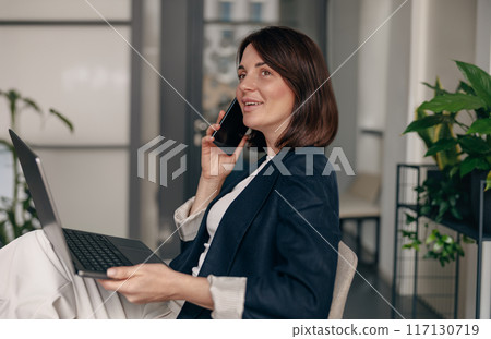 Professional businesswoman at work, smiling with phone and laptop, showing confidence and success Professional businesswoman at work, smiling with phone and laptop, showing confidence and success 117130719
