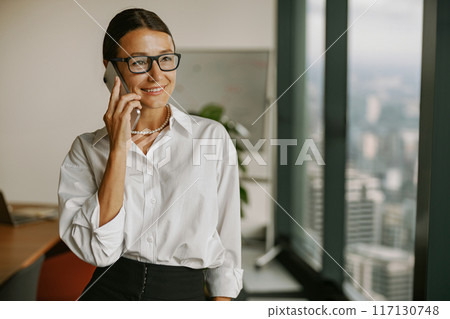 A professional businesswoman on a phone call in a modern office, showcasing confidence and ambition 117130748