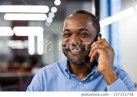 Businessman smiling and talking on phone in modern office. Expressing joy and satisfaction during a conversation. Professional setting with blurred office background. Businessman smiling and talking on phone in modern office. Expressing joy and satisfaction during a conversation. Professional setting with blurred office background. 117131736