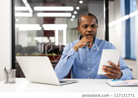 Professional man focusing intently while reading on tablet in modern office environment. Sitting at desk with laptop, thinking deeply. Professional man focusing intently while reading on tablet in modern office environment. Sitting at desk with laptop, thinking deeply. 117131771