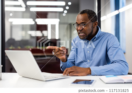 Angry African American office worker pointing finger while talking during video call at workplace. Mature businessman frustrated with laptop inside modern office setting. Angry African American office worker pointing finger while talking during video call at workplace. Mature businessman frustrated with laptop inside modern office setting. 117131825