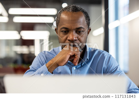 Professional businessman concentrating on tasks using laptop in modern office environment. Serious expression reflects deep thought. Ideal for themes emphasizing business, technology, productivity Professional businessman concentrating on tasks using laptop in modern office environment. Serious expression reflects deep thought. Ideal for themes emphasizing business, technology, productivity 117131843