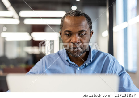 Portrait of focused office worker in blue shirt working on laptop in modern office environment. Depicts concentration, productivity, and professional setting. Suitable for business, technology, and Portrait of focused office worker in blue shirt working on laptop in modern office environment. Depicts concentration, productivity, and professional setting. Suitable for business, technology, and 117131857