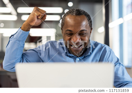 Happy businessman excitedly celebrating achievement with fist pump in modern office. Middle aged professional exuding joy and positivity, working laptop. Office environment represents accomplishment Happy businessman excitedly celebrating achievement with fist pump in modern office. Middle aged professional exuding joy and positivity, working laptop. Office environment represents accomplishment 117131859