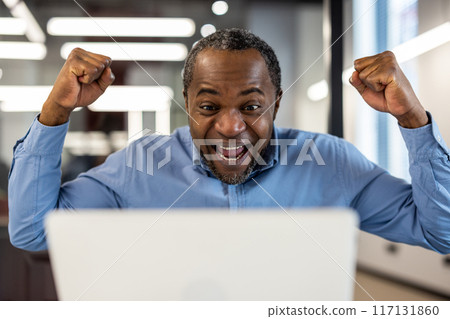Happy businessman celebrating success at work on laptop. Man with raised fists showing joy and excitement while looking at screen. Concept of achievement, victory, and career success. 117131860