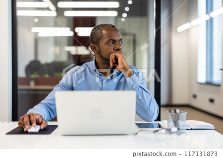 Professional businessman sitting at office desk with laptop, deep in thought. Modern workspace with natural light. Concept of business planning, strategy, and focus. Professional businessman sitting at office desk with laptop, deep in thought. Modern workspace with natural light. Concept of business planning, strategy, and focus. 117131873