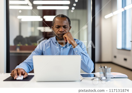 Mature african american office worker deeply focused at desk inside modern workspace. Displaying concentration and dedication while working on laptop in professional office environment. 117131874