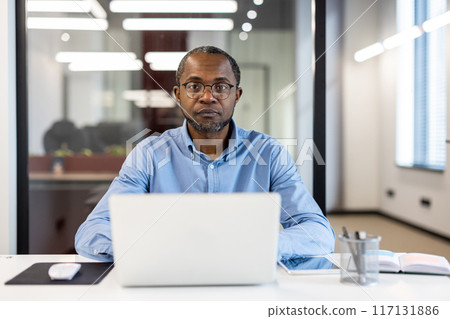 Focused professional working on laptop in modern office environment. Man wearing glasses, blue shirt, sitting at desk with notebook, mouse, and pen holder. Bright office with modern lighting. Focused professional working on laptop in modern office environment. Man wearing glasses, blue shirt, sitting at desk with notebook, mouse, and pen holder. Bright office with modern lighting. 117131886