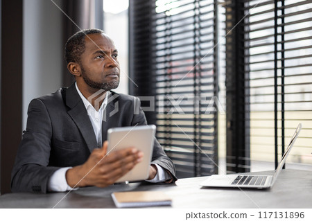 Focused African American businessman in a suit uses a digital tablet while sitting at a desk with a laptop in a contemporary office setting. Focused African American businessman in a suit uses a digital tablet while sitting at a desk with a laptop in a contemporary office setting. 117131896