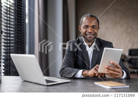A professional African American businessman engages with a digital tablet at his desk in a modern office setting, portraying confidence and expertise. 117131899
