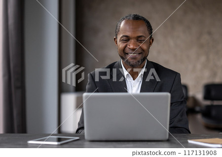 A professional African American man smiles while working on his laptop in an office setting. This image captures a moment of career satisfaction and confidence. 117131908