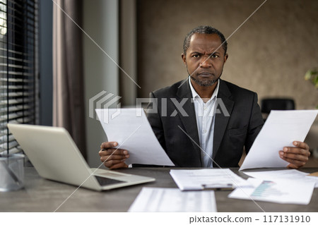 Mature African American businessman in a suit frowns as he reviews important documents, reflecting a stressful moment in a professional setting. 117131910