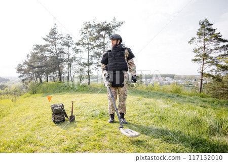 A man in a military uniform and bulletproof vest works in the forest with a metal detector. A minesweeper performs work on demining the territory A man in a military uniform and bulletproof vest works in the forest with a metal detector. A minesweeper performs work on demining the territory 117132010