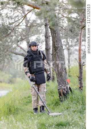A man in a military uniform and bulletproof vest works in the forest with a metal detector. A minesweeper performs work on demining the territory A man in a military uniform and bulletproof vest works in the forest with a metal detector. A minesweeper performs work on demining the territory 117132013