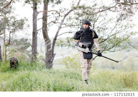 A man in a military uniform and bulletproof vest works in the forest with a metal detector. A minesweeper performs work on demining the territory 117132015