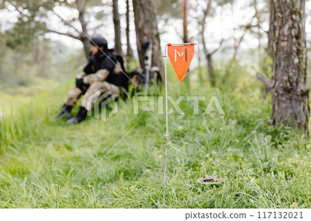 A man in a military uniform and a bulletproof vest works in the forest to demine the territory. A man warns of danger by making a red mark 117132021