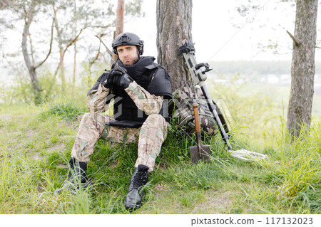 A man in a military uniform and a bulletproof vest sits in the forest near a metal detector and a military backpack. A man pauses in the work of demining the territory A man in a military uniform and a bulletproof vest sits in the forest near a metal detector and a military backpack. A man pauses in the work of demining the territory 117132023