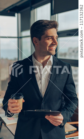 Handsome businessman in suit standing with coffee and clipboard at office on colleagues background Handsome businessman in suit standing with coffee and clipboard at office on colleagues background 117132362