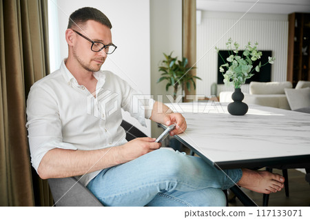 Portrait of man engaging with smartphone sitting at white marble countertop on kitchen, wearing glasses. Handsome man relaxing in his minimalist and modern aesthetic apartment. 117133071