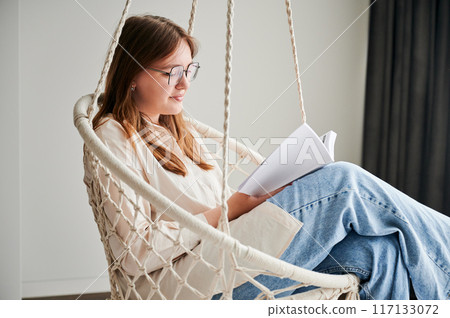 Beautiful girl relaxing and reading book on indoor swing. Pretty female in glasses excited about story she reading. Cozy afternoon with book in hanging chair in home. 117133072