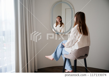 Beautiful girl looking at herself in mirror and smiling. Pretty female checking herself. Cute smiling girl sitting in chair opposite to mirror. 117133074