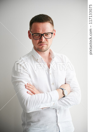 Portrait of man with serious and slightly displeased expression, upset or frustrated by something. Handsome young male in casual clothes keeping arms crossed while standing on white background. 117133075