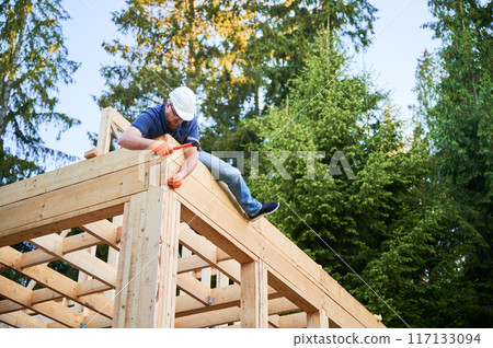 Woodworker constructing wooden, two-story frame-built house near the woods. Bearded, spectacled man driving nail into structure while wearing safety helmet. Notion of modern ecological construction. 117133094