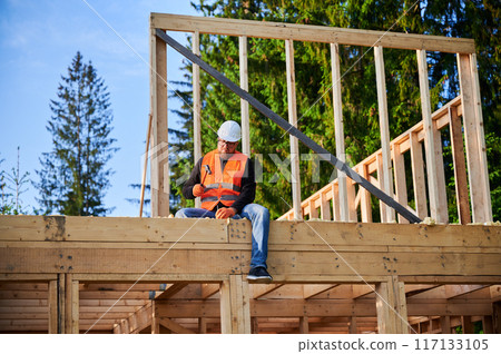Carpenter constructing two-story wooden frame house near forest. Bearded man hammering nails into the structure, wearing protective helmet, construction vest. Concept of modern ecological construction 117133105