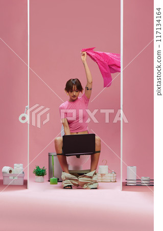 Happy, emotional young girl in pink clothes sitting in toilet and watching online game translation, supporting team against pink background Happy, emotional young girl in pink clothes sitting in toilet and watching online game translation, supporting team against pink background 117134164