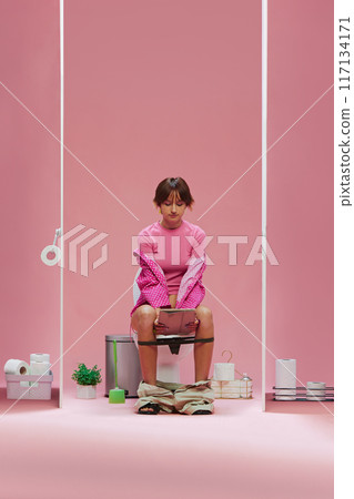 Young girl in pink clothes, with short hair sitting on toilet and looking on tablet against pink background 117134171
